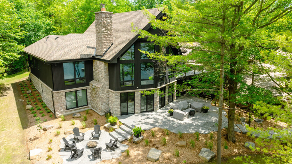 Aerial view of a modern two-story house with large glass windows and a stone facade, surrounded by greenery. The backyard features a spacious patio with outdoor seating and a fire pit, set amidst landscaped gardens and tall trees.