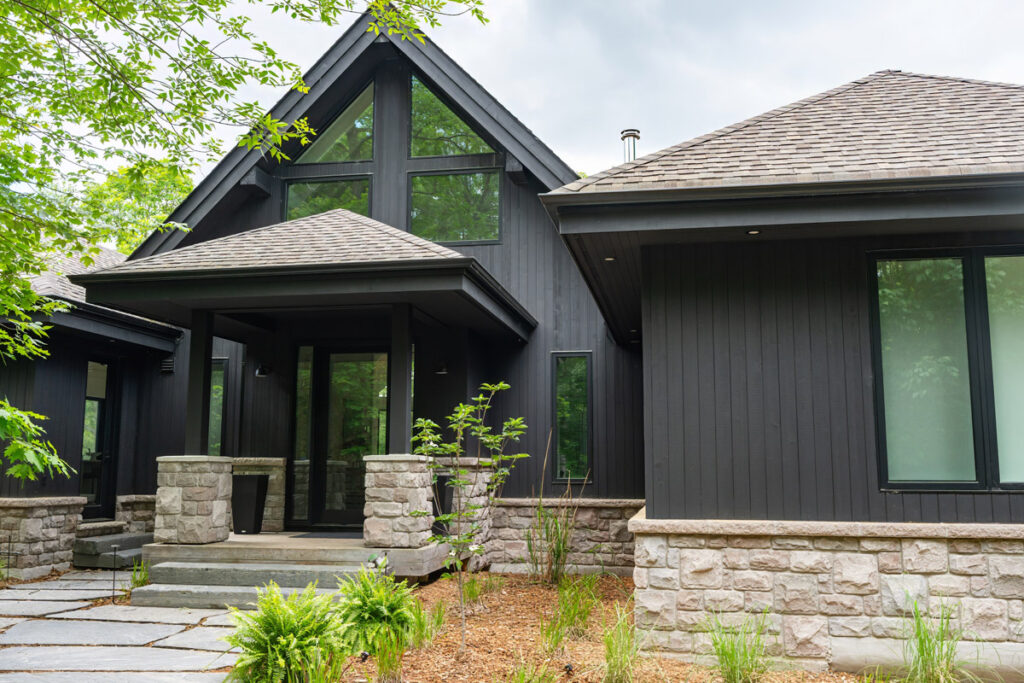 Modern black house with steep gable roof and large windows. The front features a stone pathway bordered by plants and a covered entryway supported by stone pillars. The roof is shingled, and the surrounding area is landscaped with greenery.